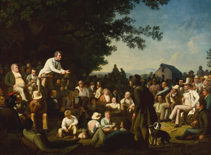 many gather around a man who stands on a tree stump to give a speech